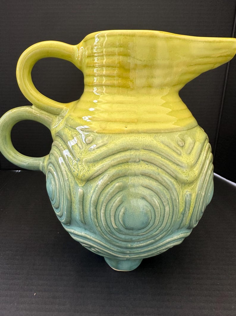 May include: A handcrafted ceramic pitcher with a yellow top and a blue-green base. The pitcher features a spout, two handles, and a textured, concentric circle design. The pitcher is approximately 9 inches tall.