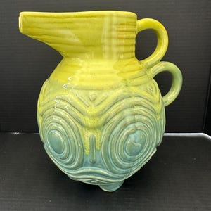May include: A ceramic pitcher with a yellow and teal color scheme. The pitcher has a spout, two handles, and a textured, patterned body. The base has three small feet. The pitcher is approximately 9 inches tall.