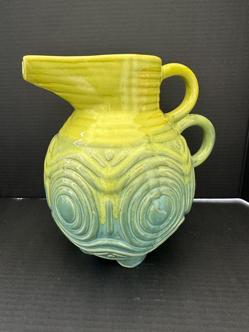 May include: A ceramic pitcher with a yellow and teal color scheme. The pitcher has a spout, two handles, and a textured, patterned body. The base has three small feet. The pitcher is approximately 9 inches tall.