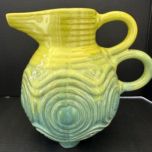 May include: A ceramic pitcher with a spout and two handles. The pitcher is a gradient of yellow and teal, with a textured, concentric circle design on the body. The pitcher has three small feet.