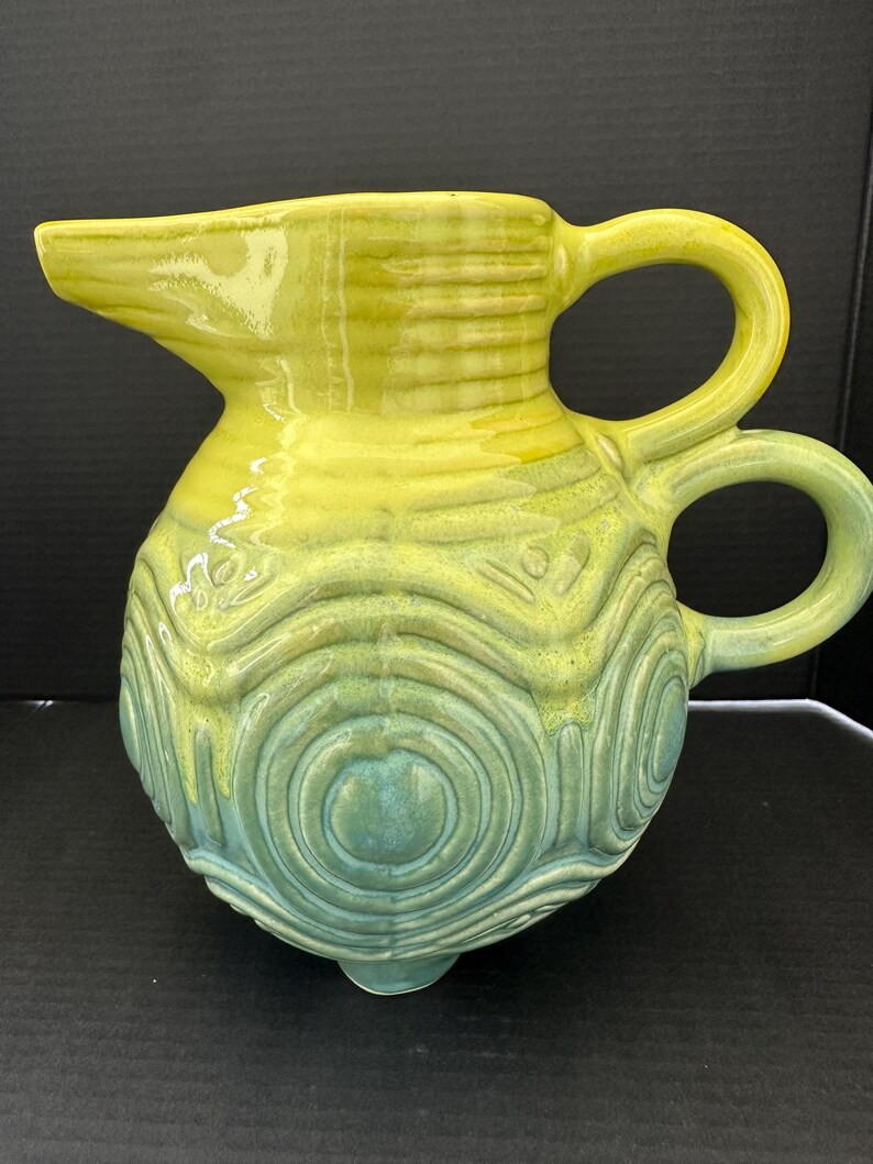 May include: A ceramic pitcher with a spout and two handles. The pitcher is a gradient of yellow and teal, with a textured, concentric circle design on the body. The pitcher has three small feet.