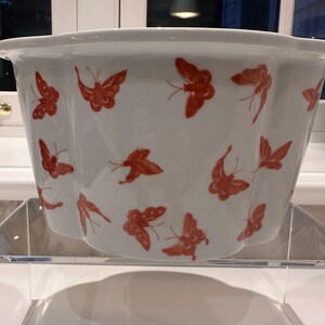 May include: A white ceramic bowl with a scalloped rim and a repeating pattern of orange butterflies printed across the exterior surface. The bowl features a deep, rounded shape with vertical indentations along the sides, creating a fluted appearance.