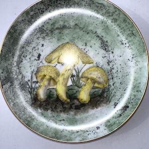 May include: A round decorative plate with a gold rim and a green and white speckled background. The plate features a hand-painted illustration of three yellow mushrooms with brown stems and green grass.