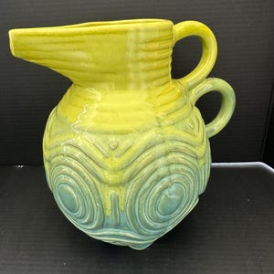 May include: A ceramic pitcher with a yellow and teal glaze. The pitcher has a spout, two handles, and a bulbous body decorated with concentric circle patterns. The pitcher is likely vintage or retro.