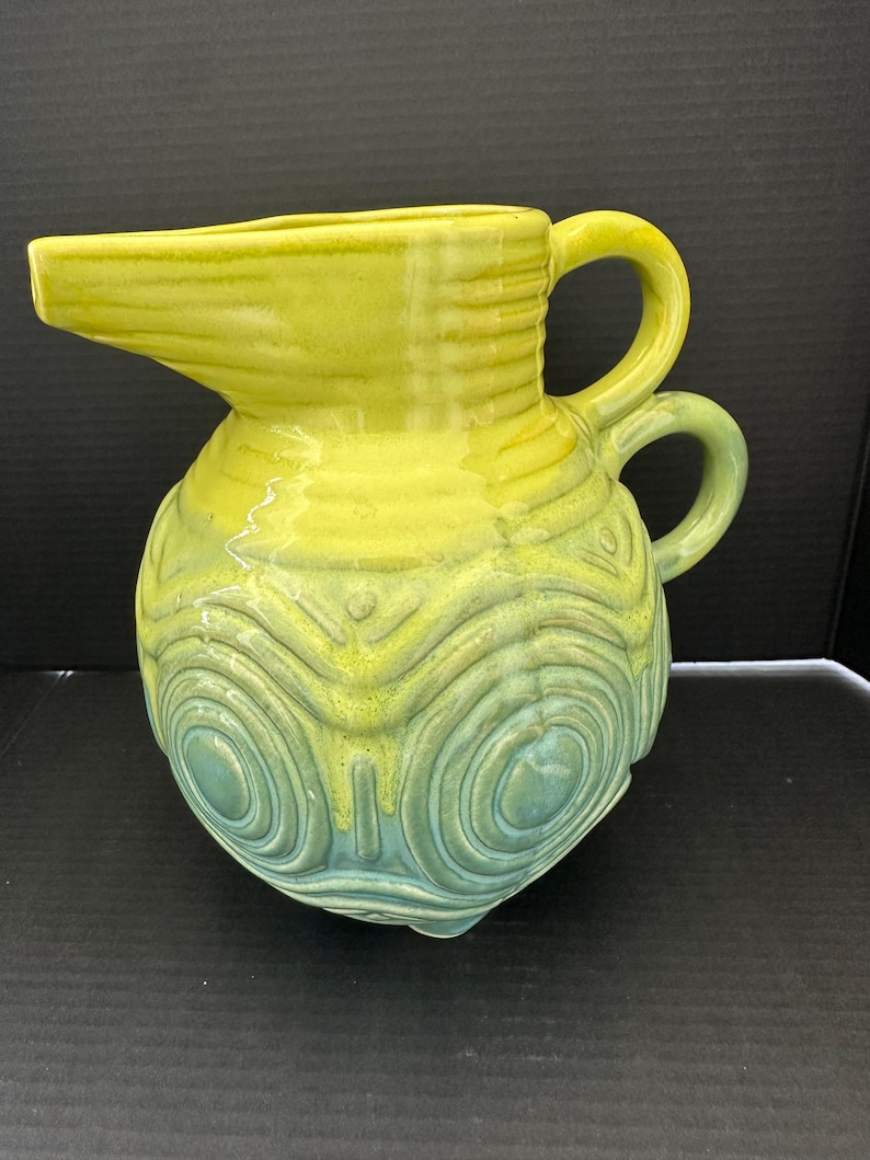 May include: A ceramic pitcher with a yellow and teal glaze. The pitcher has a spout, two handles, and a bulbous body decorated with concentric circle patterns. The pitcher is likely vintage or retro.