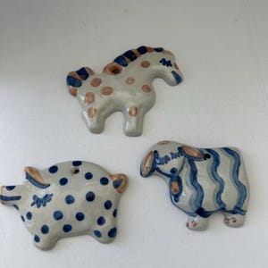 May include: Three ceramic animal figurines: a horse with brown spots and blue accents, a pig with blue polka dots, and a sheep with blue stripes. Each has a hole for hanging. The figurines are white with blue and brown details.