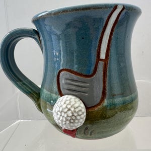 May include: A ceramic mug with a blue and green glaze. The mug features a golf club and golf ball design. The golf ball is on a red tee. The mug has a curved handle.