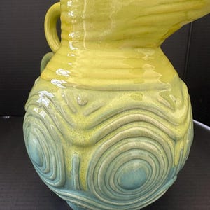 May include: A ceramic pitcher with a yellow and teal glaze. The pitcher has a handle and a spout. The lower portion features a textured, concentric circle design. The pitcher sits on three small feet.