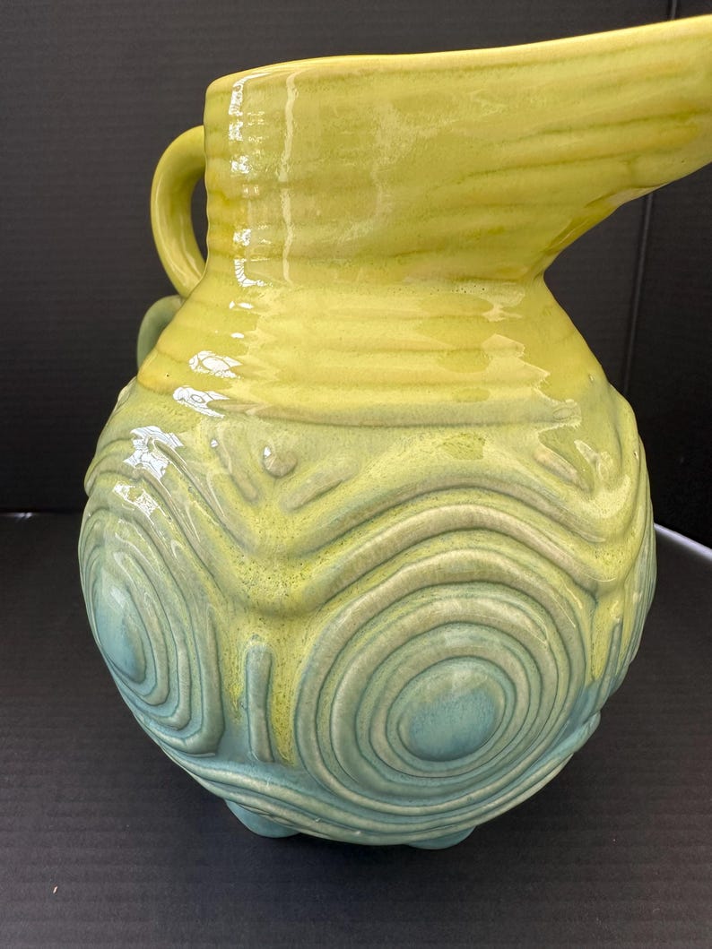 May include: A ceramic pitcher with a yellow and teal glaze. The pitcher has a handle and a spout. The lower portion features a textured, concentric circle design. The pitcher sits on three small feet.