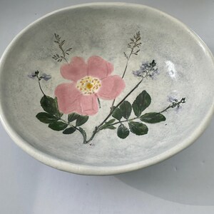 May include: A ceramic bowl with a light gray speckled finish. The bowl is decorated with a pink flower, green leaves, and sprigs of purple and green foliage. The bowl has a wide, shallow shape.