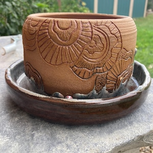 May include: A brown ceramic planter with a detailed, carved design. The planter sits on a matching saucer. The design features repeating patterns and textures. The planter is round and has a wide opening.
