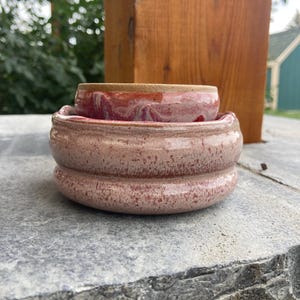 May include: A handmade ceramic pottery set in shades of pink and red. The set includes a small bowl nested inside a larger, tiered bowl. The pottery has a speckled texture and a glossy finish, perfect for a decorative accent.