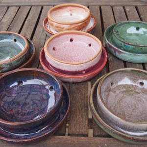 Op de afbeelding: Een verzameling handgemaakte keramische kommen in diverse kleuren, waaronder blauw, groen, roze en bruin. Elke kom heeft een bijpassende schotel en drie afvoergaten. De kommen zijn gerangschikt op een houten oppervlak, wat hun unieke texturen en glazuren laat zien.