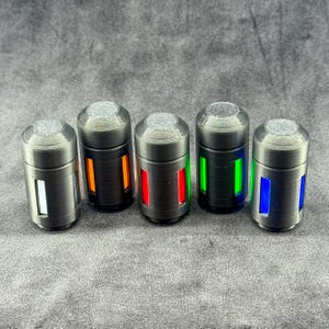 May include: Five cylindrical, metallic-colored objects with a textured top and a rectangular light-up section. The lights are white, orange, red, green, and blue. The objects are arranged in a row against a gray background.