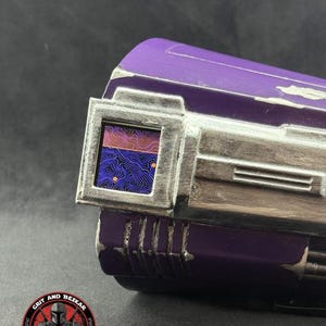 May include: Close-up of a purple and silver cosplay prop with a rectangular window displaying a colorful pattern. The prop has a weathered appearance and the text "GRIT AND BESKAR" is visible.