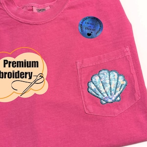Puede incluir: Camiseta rosa con bolsillo bordado con un diseño de concha marina azul y blanco. La camiseta tiene las palabras "Premium Embroidery" y "Thank you so much for your order".