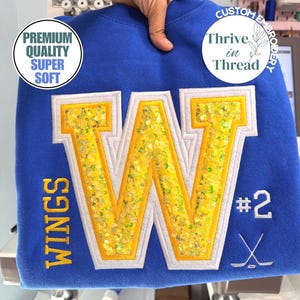 May include: A royal blue sweatshirt featuring a large central letter W with yellow sequin fill and white trim. The text WINGS is embroidered in yellow on the left, while #2 and a crossed hockey stick graphic are embroidered in white on the right.
