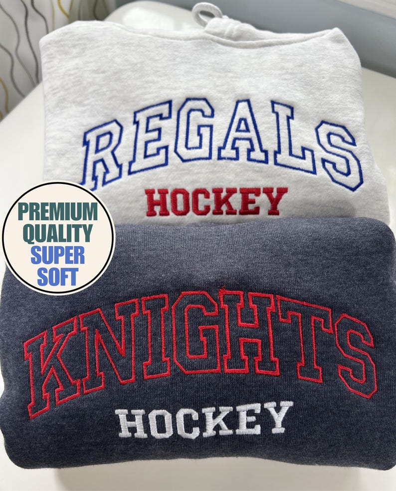 Puede incluir: Dos sudaderas dobladas, una gris claro y otra gris oscuro. La sudadera gris claro tiene bordado "REGALS" en azul y "HOCKEY" en rojo. La sudadera gris oscuro tiene bordado "KNIGHTS" en rojo y "HOCKEY" en blanco. Un gr&aacute;fico circular dice "PREMIUM QUALITY SUPER SOFT".