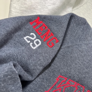 Pu&ograve; includere: Primo piano di una felpa con cappuccio grigio scuro con testo ricamato rosso che dice "MENG" e il numero "29". La parola "KING" &egrave; ricamata in rosso. Una felpa con cappuccio grigio chiaro &egrave; visibile sullo sfondo.