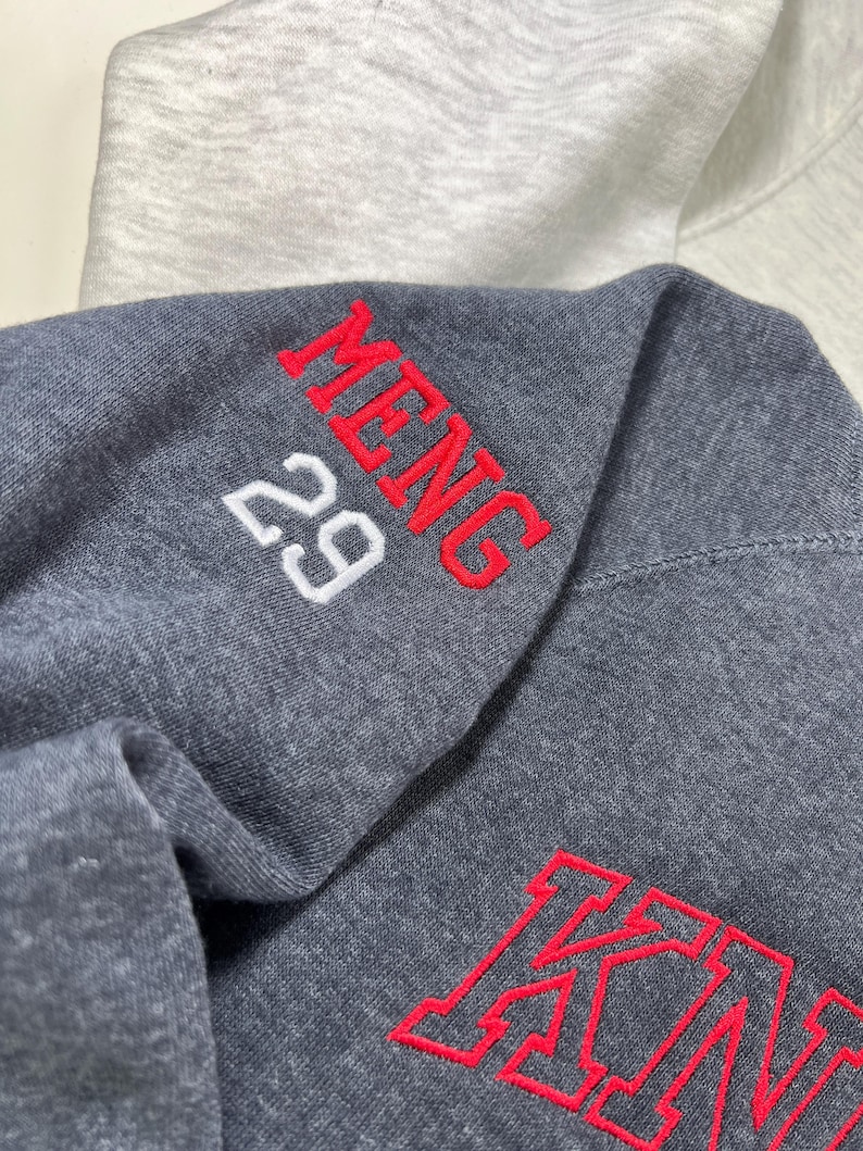 Pu&ograve; includere: Primo piano di una felpa con cappuccio grigio scuro con testo ricamato rosso che dice "MENG" e il numero "29". La parola "KING" &egrave; ricamata in rosso. Una felpa con cappuccio grigio chiaro &egrave; visibile sullo sfondo.
