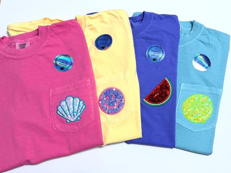 May include: Four short-sleeved t-shirts in pink, yellow, purple, and light blue, each featuring a chest pocket with a unique patch: a seashell, a smiley face, a watermelon slice, and a citrus slice. Each shirt includes a circular holographic sticker.