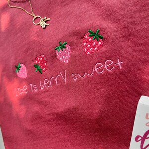May include: A red-pink t-shirt with embroidered strawberries and the phrase "life is berry sweet" in pink thread. A gold necklace with a small charm is visible near the top. The shirt has a soft, textured appearance.