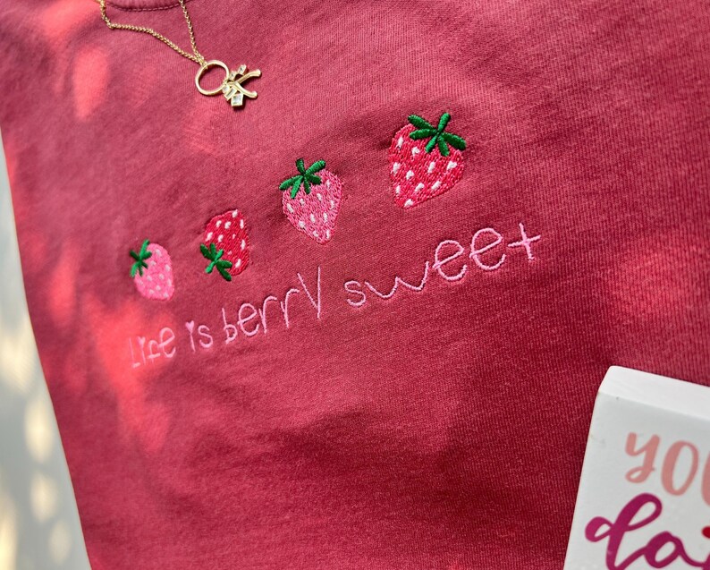 May include: A red-pink t-shirt with embroidered strawberries and the phrase "life is berry sweet" in pink thread. A gold necklace with a small charm is visible near the top. The shirt has a soft, textured appearance.