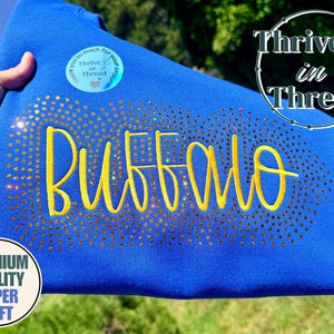 Pode incluir: Uma camisola azul royal com a palavra BUFFALO em letras amarelas. O texto é rodeado por uma borda retangular densa de strass dourados cintilantes. Um autocolante circular com o texto Thrive in Thread é visível.