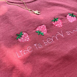 May include: A dusty rose-colored t-shirt with embroidered strawberries and the phrase "Life is berry sweet." A gold necklace with a leaf pendant is draped across the shirt.