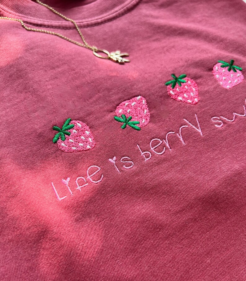 May include: A dusty rose-colored t-shirt with embroidered strawberries and the phrase "Life is berry sweet." A gold necklace with a leaf pendant is draped across the shirt.