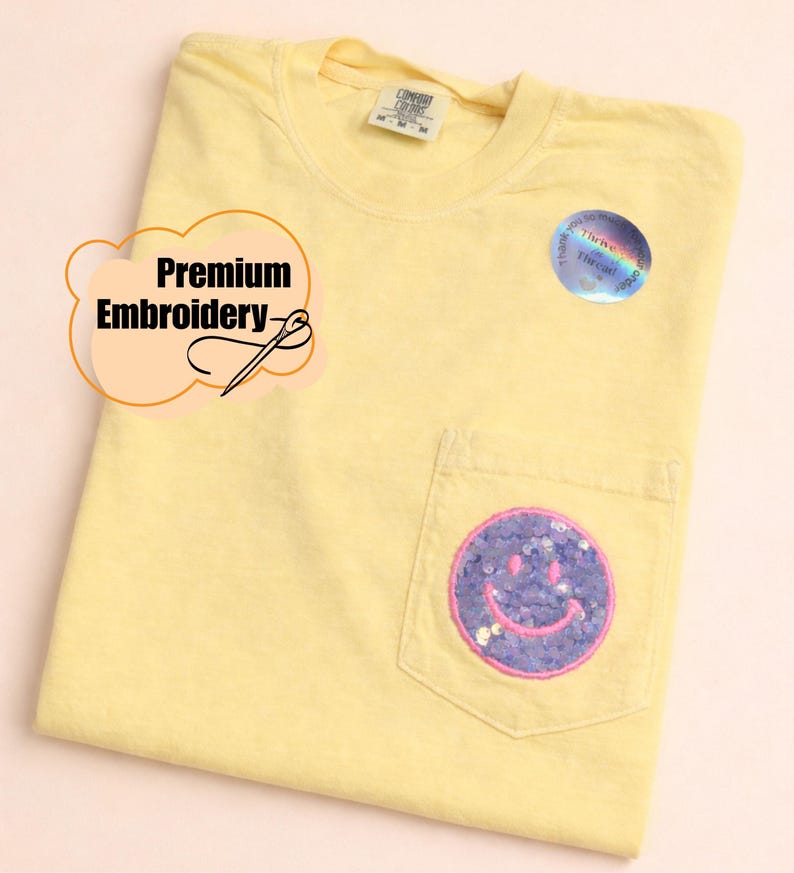 May include: A pale yellow short-sleeve t-shirt with a chest pocket featuring a circular purple sequin smiley face patch with pink stitching. The shirt has a Comfort Colors label at the neck and a holographic sticker. Text reads Premium Embroidery.