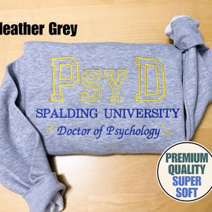 May include: Heather grey sweatshirt with "Psy D" in yellow embroidery. Below, the text reads "Spalding University Doctor of Psychology" in blue. A circular graphic states "Premium Quality Super Soft".