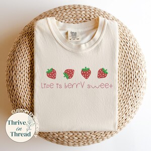 May include: Cream-colored t-shirt with a crew neck, featuring four embroidered strawberries in a row. The phrase "Life is berry sweet" is embroidered below the strawberries in a cursive font. The shirt is displayed on a woven, circular surface.