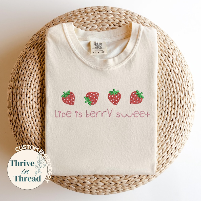 May include: Cream-colored t-shirt with a crew neck, featuring four embroidered strawberries in a row. The phrase "Life is berry sweet" is embroidered below the strawberries in a cursive font. The shirt is displayed on a woven, circular surface.