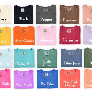 May include: A collection of twenty-eight short-sleeve t-shirts arranged in a grid, each featuring a neck label and a unique color name printed in white text, including White, Ivory, Black, Pepper, Espresso, Peachy, Butter, Watermelon, Citrus, Burnt Orange, Blossom, Crimson, Orchid, Bay, Violet, Chambray, Chalky Mint, Light Green, Blue Jean, Moss, Blue Spruce, Neon Lemon, Neon Pink, Neon Violet, Flo Blue, Neon Red Orange, Neon Cantaloupe, and Heliconia.