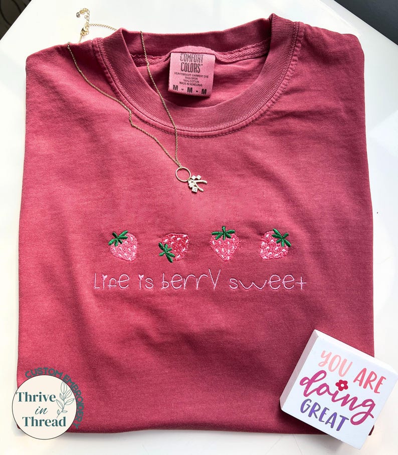 May include: A maroon Comfort Colors t-shirt with embroidered strawberries and the phrase "Life is berry sweet." A gold necklace and a small wooden block with the text "YOU ARE doing GREAT" are also present.