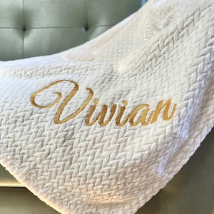 May include: A white, textured blanket with a woven pattern. The name "Vivian" is embroidered in gold thread across the blanket. The blanket is draped over a green upholstered chair.