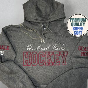 Puede incluir: Una sudadera con capucha gris con un collar dorado. La parte delantera de la sudadera tiene las palabras "Orchard Park HOCKEY" bordadas en rojo y blanco. La manga tiene "GOALIE MOM" bordado en rojo. La imagen también tiene el texto "PREMIUM QUALITY SUPER SOFT".