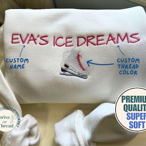 以下が含まれることがあります： ピンク色の糸で「EVA'S ICE DREAMS」と刺繍された白いスウェットシャツ。テキストの下にはフィギュアスケートブーツが刺繍されています。「Premium Quality Super Soft」のラベルが付いています。