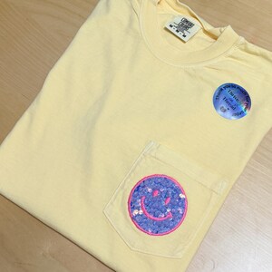 May include: A pale yellow short-sleeve t-shirt with a chest pocket featuring a circular patch with purple sequins and a pink embroidered smiley face. The shirt has a Comfort Colors label and a round sticker that reads Thank you so much for your order.