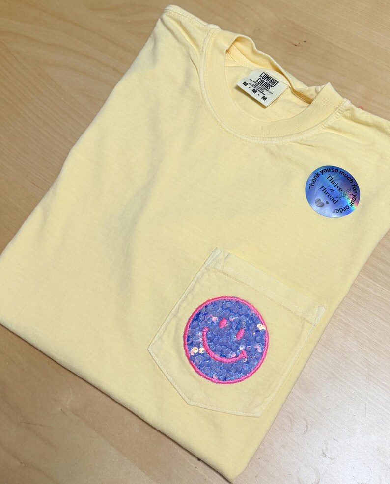 May include: A pale yellow short-sleeve t-shirt with a chest pocket featuring a circular patch with purple sequins and a pink embroidered smiley face. The shirt has a Comfort Colors label and a round sticker that reads Thank you so much for your order.