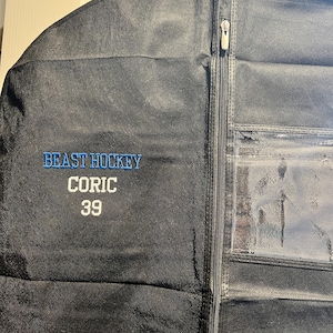 Peut inclure: Housse de vêtement noire avec le texte "BEAST HOCKEY" en bleu, "CORIC" en blanc et le numéro "39" en blanc. Le sac comporte une fenêtre en plastique transparent et une fermeture éclair sur toute la longueur.