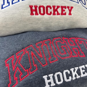 Pu&ograve; includere: Primo piano di due felpe. La felpa superiore &egrave; grigio chiaro con testo ricamato blu e rosso che dice "REGALS HOCKEY". La felpa inferiore &egrave; grigio scuro con testo ricamato rosso e bianco che dice "KNIGHT HOCKEY".