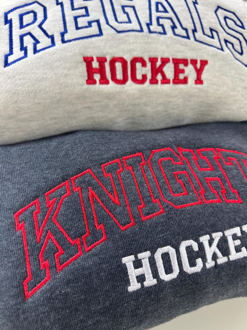 Pu&ograve; includere: Primo piano di due felpe. La felpa superiore &egrave; grigio chiaro con testo ricamato blu e rosso che dice "REGALS HOCKEY". La felpa inferiore &egrave; grigio scuro con testo ricamato rosso e bianco che dice "KNIGHT HOCKEY".