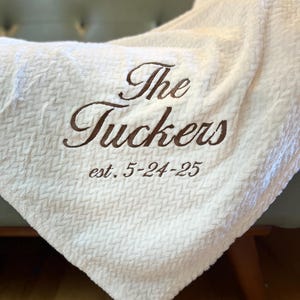 Puede incluir: Una manta blanca texturizada con el texto bordado "The Tuckers" en escritura marrón oscuro. Debajo, se lee "est. 5-24-25". La manta está sobre un sofá verde claro.