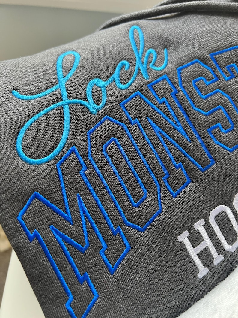 Pu&ograve; includere: Primo piano di una felpa con cappuccio grigia con le parole "Lock Monsters" ricamate con filo blu e bianco. La parola "Lock" &egrave; in corsivo, mentre "Monsters" &egrave; in stampatello. La felpa ha un cappuccio con coulisse.