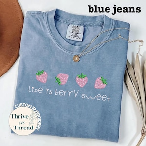May include: A light blue t-shirt with embroidered pink strawberries and the phrase "Life is berry sweet." A gold necklace with a pendant rests on the shirt. The shirt has a Comfort Colors tag and the words "blue jeans" are at the top.