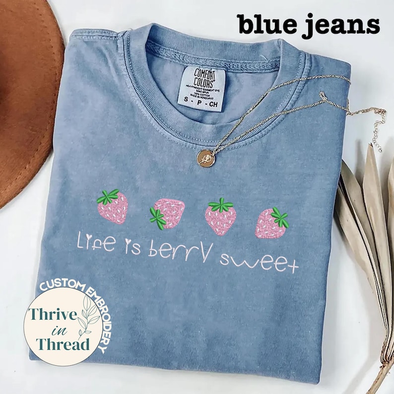 May include: A light blue t-shirt with embroidered pink strawberries and the phrase "Life is berry sweet." A gold necklace with a pendant rests on the shirt. The shirt has a Comfort Colors tag and the words "blue jeans" are at the top.