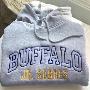 Puede incluir: Una sudadera con capucha gris con la palabra "BUFFALO" en letras azules y blancas, y "JR. SABRES" en letras doradas. La sudadera tiene un cord&oacute;n y una capucha.