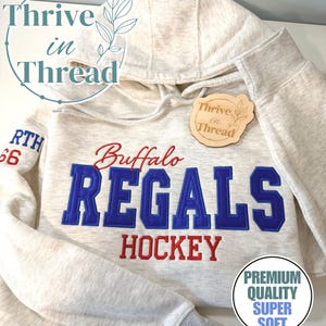 May include: A heathered oatmeal-colored hooded sweatshirt featuring embroidered text that reads Buffalo REGALS HOCKEY in blue and red thread. The sleeve displays the text NORTH 66 in blue and red. A wooden tag with the text Thrive in Thread is attached.